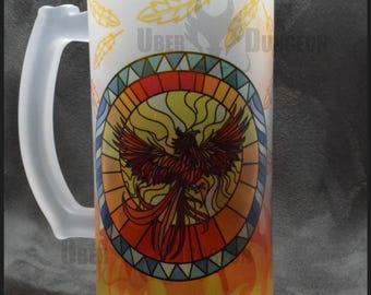 May include: A frosted glass beer mug with a handle. The mug features a colorful stained glass design of a phoenix rising from flames. The design is surrounded by a circle of yellow feathers.