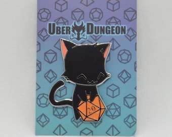 May include: Black cat enamel pin holding an orange D20 die. The pin has a silver outline and pink accents on the ears. The background is a gradient of blue and purple with geometric shapes and the text "UBER DUNGEON".