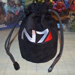 Puede incluir: Bolsa negra con cordón y el logotipo N7 bordado en blanco, plateado y rojo. La bolsa está hecha de un material suave y oscuro y tiene un cierre de cordón. Ideal para guardar objetos pequeños.