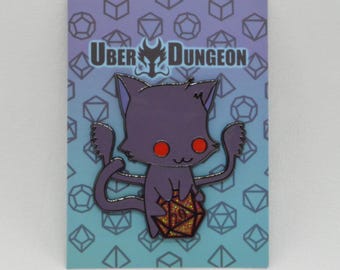 May include: A purple cat enamel pin holding a glittery gold twenty-sided die. The pin has red eyes and is set against a blue and purple background with geometric shapes. The text "UBER DUNGEON" is at the top.