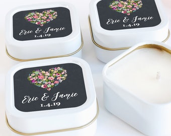 May include: Four white square candles in metal tins. Each tin has a gold rim and a black label with a floral heart design and the text "Eric & Jamie 1.4.19". One candle is open, revealing the white wax and wick.