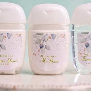 May include: Three small, white hand sanitizer bottles with white flip-top lids. Each bottle has a label with a floral design in green, gold, and blue. Text on the labels reads "Welcome to Lorenas Shower 12.22.21", "Mr. & Mrs. McKensie", and "Welcome Baby Thomas Anthony due June 2022".