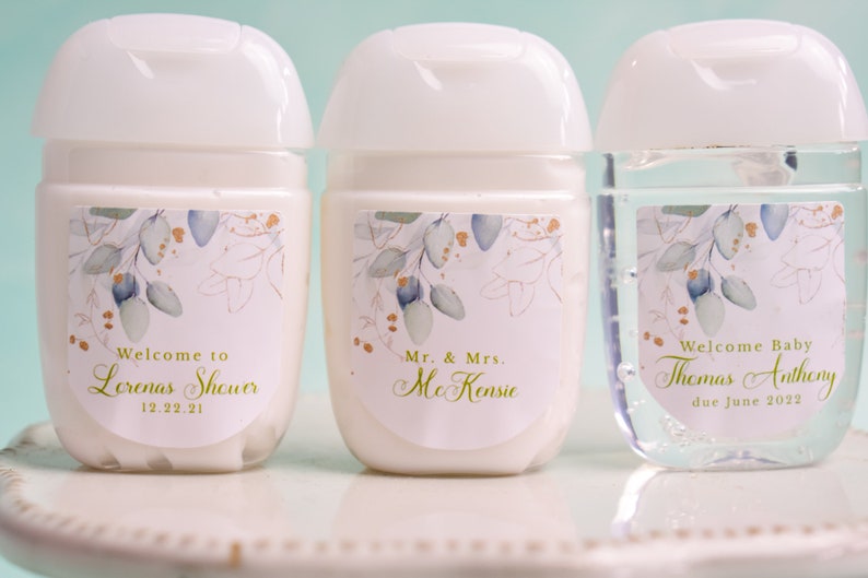 May include: Three small, white hand sanitizer bottles with white flip-top lids. Each bottle has a label with a floral design in green, gold, and blue. Text on the labels reads "Welcome to Lorenas Shower 12.22.21", "Mr. & Mrs. McKensie", and "Welcome Baby Thomas Anthony due June 2022".