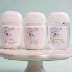 May include: Three white hand sanitizer bottles with labels. The labels feature floral designs and text that reads "Welcome to Lorenas Shower 12.22.21", "Mr. & Mrs. McKenzie", and "Welcome Baby Thomas Anthony due June 2022".