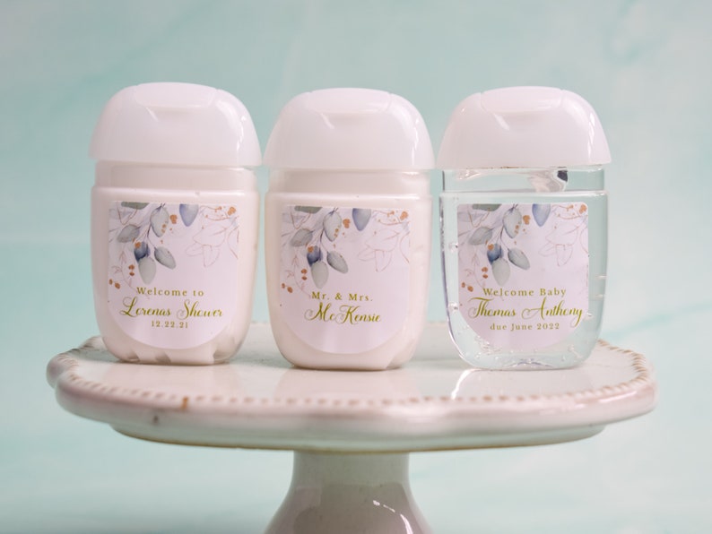 May include: Three white hand sanitizer bottles with labels. The labels feature floral designs and text that reads "Welcome to Lorenas Shower 12.22.21", "Mr. & Mrs. McKenzie", and "Welcome Baby Thomas Anthony due June 2022".