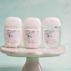 May include: Three white hand sanitizer bottles with floral labels. The labels read "Welcome to Lermas Sheurs", "Mr. & Mrs. McKense", and "Welcome Baby Thomas Anthony". The bottles sit on a white cake stand.