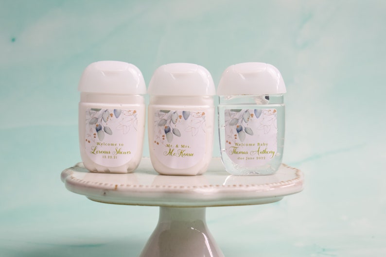 May include: Three white hand sanitizer bottles with floral labels. The labels read "Welcome to Lermas Sheurs", "Mr. & Mrs. McKense", and "Welcome Baby Thomas Anthony". The bottles sit on a white cake stand.