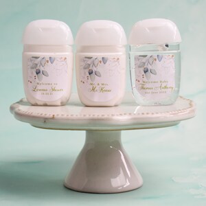 May include: Three white and clear hand sanitizer bottles with white lids, each with a personalized label. The labels feature a floral design with gold accents and text that reads "Welcome to", "Mr. & Mrs.", and "Welcome Baby". The bottles are displayed on a white cake stand.
