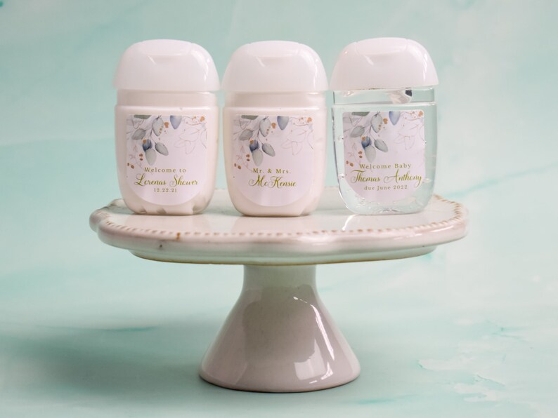 May include: Three white and clear hand sanitizer bottles with white lids, each with a personalized label. The labels feature a floral design with gold accents and text that reads "Welcome to", "Mr. & Mrs.", and "Welcome Baby". The bottles are displayed on a white cake stand.