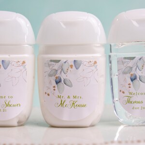 May include: Three small, white hand sanitizer bottles with white flip-top lids. Each bottle has a label with a floral design in blue, gold, and green. The labels include text for a baby shower and a wedding.
