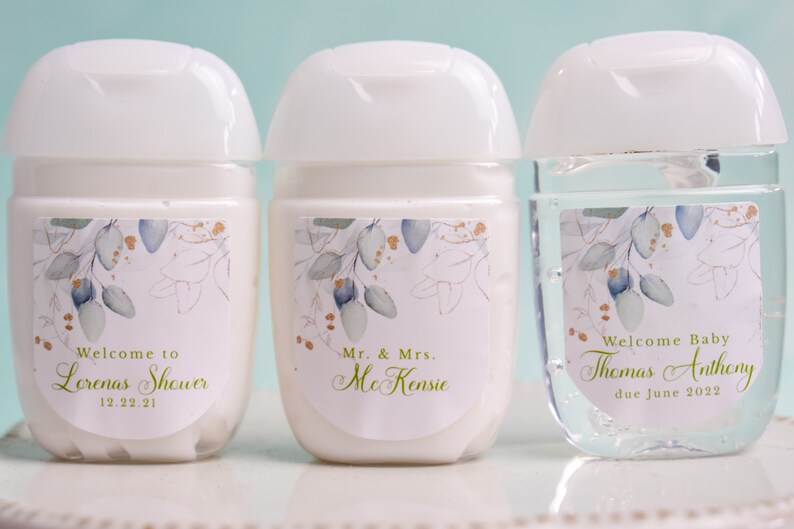 May include: Three small, white hand sanitizer bottles with white flip-top lids. Each bottle has a label with a floral design in blue, gold, and green. The labels include text for a baby shower and a wedding.