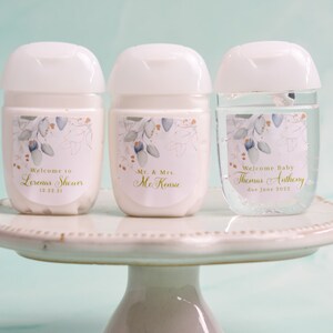 May include: Three white hand sanitizer bottles with floral labels and white caps are displayed on a white cake stand. The labels read "Welcome to Lorena's Shower 12.23.21", "Mr. & Mrs. McKenzie", and "Welcome Baby Thomas Anthony due June 2022".
