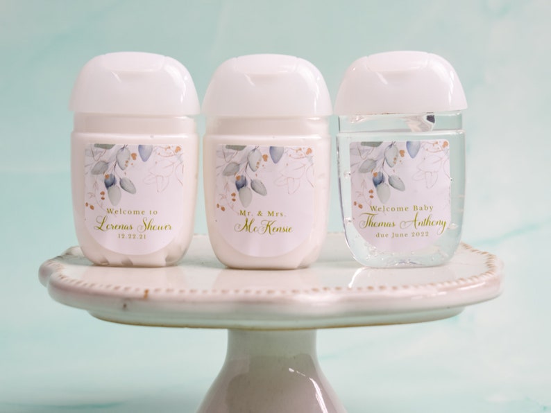 May include: Three white hand sanitizer bottles with floral labels and white caps are displayed on a white cake stand. The labels read "Welcome to Lorena's Shower 12.23.21", "Mr. & Mrs. McKenzie", and "Welcome Baby Thomas Anthony due June 2022".