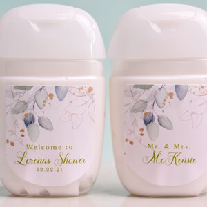 May include: Two white, cylindrical hand sanitizer bottles with white flip-top lids. Each bottle has a label with a floral design in gold, blue, and green. One label reads "Welcome to Lorenas Shower 12.22.21" and the other reads "Mr. & Mrs. McKenzie."