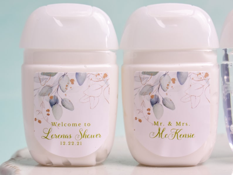 May include: Two white, cylindrical hand sanitizer bottles with white flip-top lids. Each bottle has a label with a floral design in gold, blue, and green. One label reads "Welcome to Lorenas Shower 12.22.21" and the other reads "Mr. & Mrs. McKenzie."