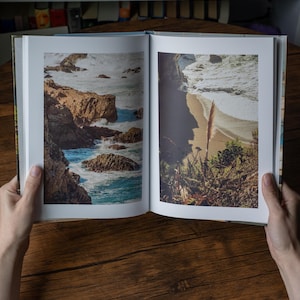 May include: An open hardcover book displaying two full-page color photographs of a coastal landscape. The left page shows rocky cliffs meeting crashing ocean waves, while the right page depicts a sandy beach with vegetation and surf.