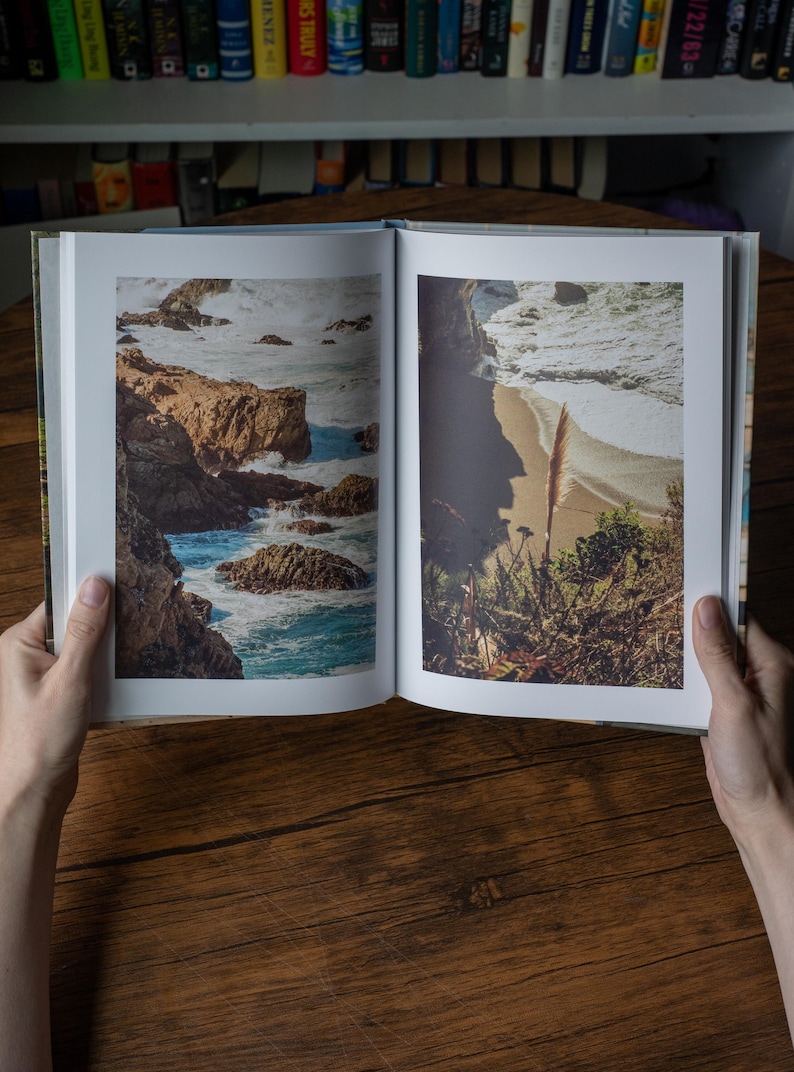 May include: An open hardcover book displaying two full-page color photographs of a coastal landscape. The left page shows rocky cliffs meeting crashing ocean waves, while the right page depicts a sandy beach with vegetation and surf.