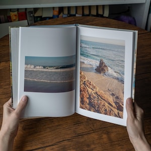 May include: An open hardcover book displaying two distinct color photographs of a coastal scene. The left page shows a horizontal view of ocean waves, while the right page features a vertical image of a rocky shoreline with sand and sea.