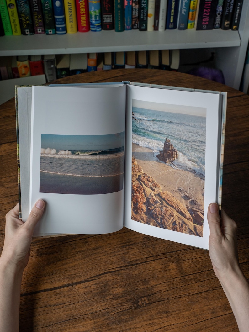 May include: An open hardcover book displaying two distinct color photographs of a coastal scene. The left page shows a horizontal view of ocean waves, while the right page features a vertical image of a rocky shoreline with sand and sea.