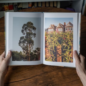 May include: An open hardcover book displaying two distinct photographs on its pages. The left page features a tall, slender eucalyptus tree against a clear sky. The right page shows a row of suburban houses partially obscured by green foliage.