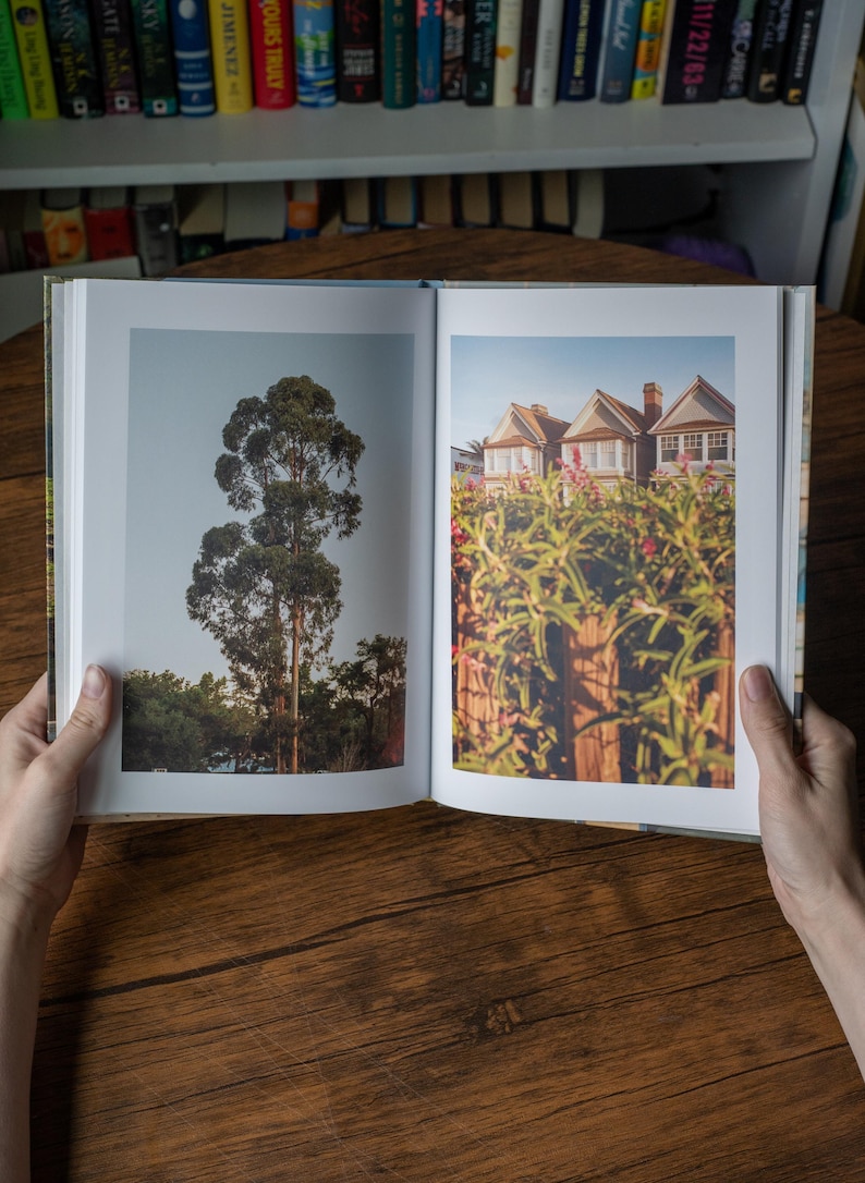 May include: An open hardcover book displaying two distinct photographs on its pages. The left page features a tall, slender eucalyptus tree against a clear sky. The right page shows a row of suburban houses partially obscured by green foliage.