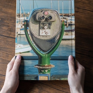 May include: A hardcover book featuring a photograph of a green metal coin-operated public viewing binocular stand. The device has two eyepieces, a coin slot labeled Quarters Only, and a small graphic of a bird. The background shows a marina.
