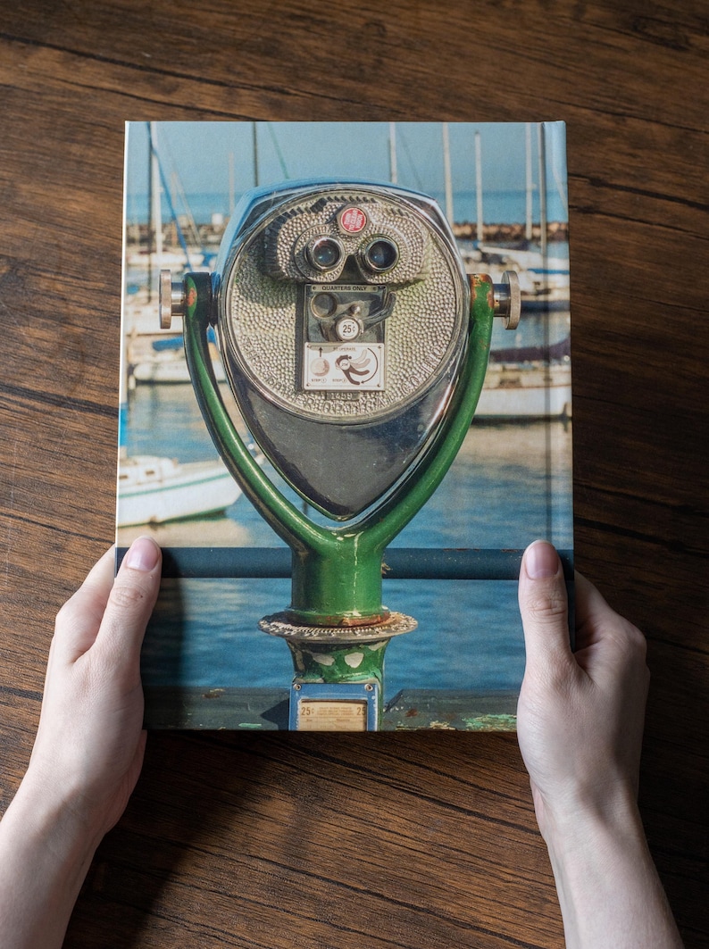 May include: A hardcover book featuring a photograph of a green metal coin-operated public viewing binocular stand. The device has two eyepieces, a coin slot labeled Quarters Only, and a small graphic of a bird. The background shows a marina.