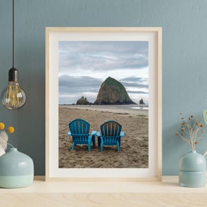 Op de afbeelding: Een ingelijste foto met twee blauwe Adirondack-stoelen op een zandstrand. Op de achtergrond rijst een grote rotsformatie op uit de oceaan onder een bewolkte hemel. De afbeelding staat in een licht houten frame.