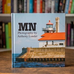 May include: A hardcover book titled MN Photography by Anthony Londer featuring a cover image of a white lighthouse with an orange roof and a tall chimney, situated on a concrete pier extending into blue water under a clear sky.