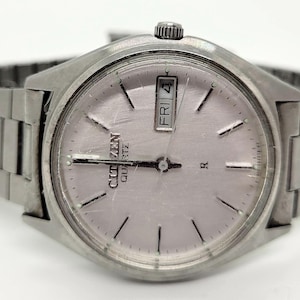 May include: Silver-toned Citizen Quartz watch with a light pink face. The watch features a day and date display and a silver metal band. The watch face has black hour markers and hands. The watch is a classic style.