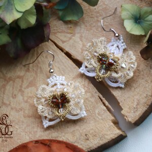 Pode incluir: Um par de brincos brancos e dourados com um detalhe de cristal castanho e transparente. Os brincos são feitos de renda e contas delicadas.