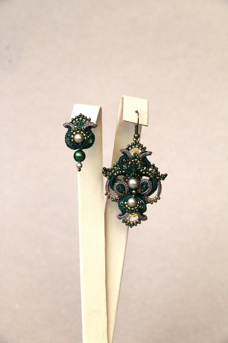 May include: A pair of green and silver earrings with intricate designs and pearl accents. The earrings are displayed on white stands.