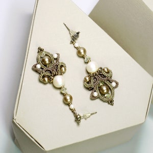 May include: A pair of dangle earrings with a delicate, intricate design. The earrings feature a combination of small, white pearls, gold beads, and a brown crocheted pattern. The earrings are displayed on a white, triangular box.