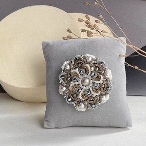 Puede incluir: Un cojín de alfileres de terciopelo gris con una gran broche de flor de cuentas plateadas y doradas. La broche tiene un centro de perla y está rodeada de cuentas trabajadas.