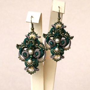 May include: A pair of green and gray beaded earrings with a pearl accent. The earrings are displayed on a white stand.