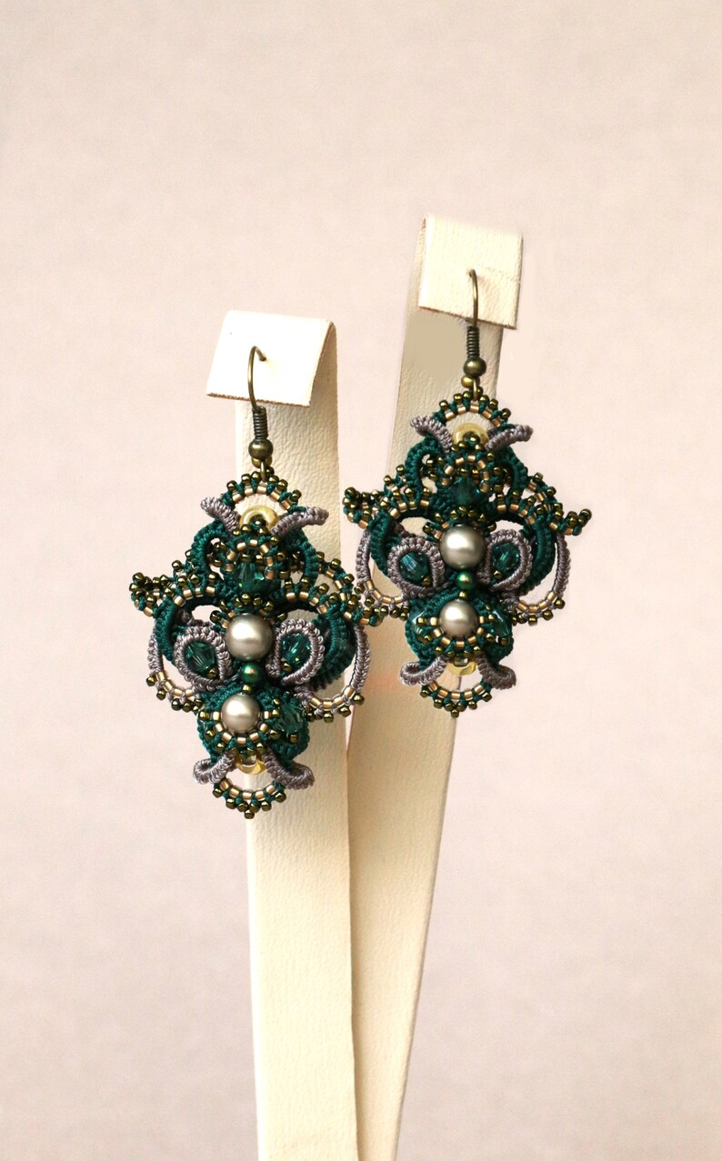 May include: A pair of green and gray beaded earrings with a pearl accent. The earrings are displayed on a white stand.