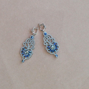 Może przedstawiać: Para srebrnych kolczyków wiszących z niebieskimi kryształowymi akcentami. Kolczyki mają delikatny, misterny wzór.