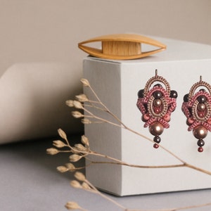 Könnte beinhalten: Ein Paar rosa und braune Perlenohrringe mit einem zarten, filigranen Design. Die Ohrringe werden auf einer weißen Schachtel mit einer Holzspule präsentiert.
