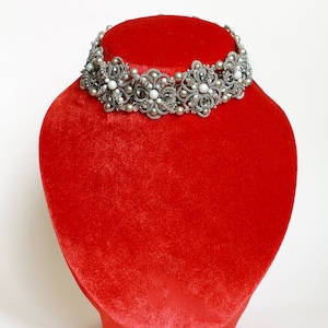 Puede incluir: Un collar gargantilla de cuentas plateadas y blancas con un diseño floral, exhibido en un busto de terciopelo rojo.