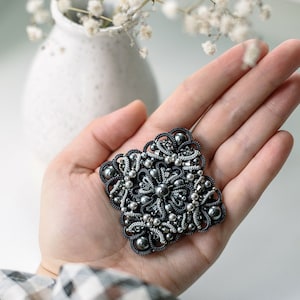 以下が含まれることがあります： 黒と銀のデザインの正方形のブローチ。ブローチは複雑なワイヤーワークで作られており、小さなビーズとパールが特徴です。