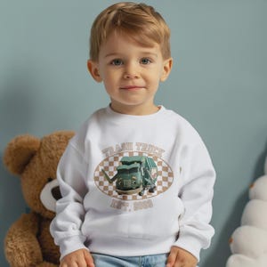 Puede incluir: Sudadera blanca para niños con un diseño de camión de basura de dibujos animados dentro de un óvalo a cuadros marrón y blanco. El texto "TRASH TRUCK" está encima del camión, con "EST. 2020" debajo.