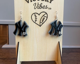 May include: Black and gray acrylic earrings in the shape of the New York Yankees logo, with the numbers 22 and 99. The earrings hang from silver hooks and are displayed on a wooden stand with the text "VICTORY Vibes co" and a heart-shaped baseball and football design.
