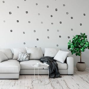 Op de afbeelding: Een moderne woonkamer met een witte hoekbank, kussens en een kleine, ronde bijzettafel. Een donkergrijze deken is over de bank gedrapeerd. De witte muur is versierd met kleine, vierkante, grijze en zwarte vormen. Een potplant staat in de hoek.