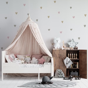 Może przedstawiać: Pokój dziecięcy z białym łóżkiem, baldachimem i dekoracyjnymi poduszkami. Drewniana szafka mieści zabawki i światło w kształcie gwiazdy. Szary dywan w kształcie liścia i pluszowa zabawka w kształcie wieloryba leżą na podłodze. Ściany zdobią serca.