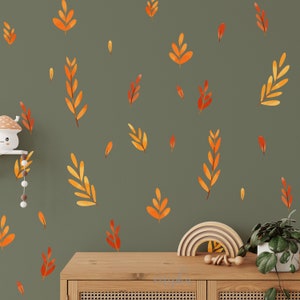 Op de afbeelding: Een muur versierd met oranje en gele bladstickers. De bladeren zijn in verschillende maten en vormen verspreid over een gedempte groene achtergrond. Een houten dressoir en decoratieve items staan op de voorgrond.