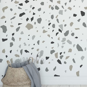 May include: A woven basket with handles, filled with a gray blanket, sits on a white wooden floor. The wall behind the basket is painted white and decorated with a terrazzo pattern in shades of gray, black, and beige.
