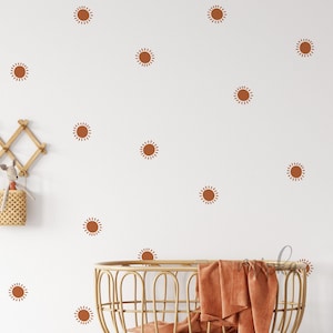 Może przedstawiać: Biała ściana z łóżeczkiem i zestawem 15 naklejek ściennych w kształcie słońca w kolorze terakoty. Naklejki są rozmieszczone na ścianie w losowym wzorze.