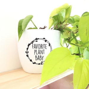 Pode incluir: Vaso de cerâmica branco com letras pretas que dizem "Favorite Plant Baby" em um círculo com um design de coroa. Uma planta verde cresce fora do vaso.
