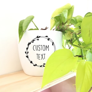 Può includere: Vaso in ceramica bianca con un design a corona nera e il testo "CUSTOM TEXT". Una pianta verde sta crescendo nel vaso.