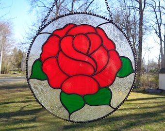 May include: A stained glass window hanging with a red rose with green leaves on a white background.